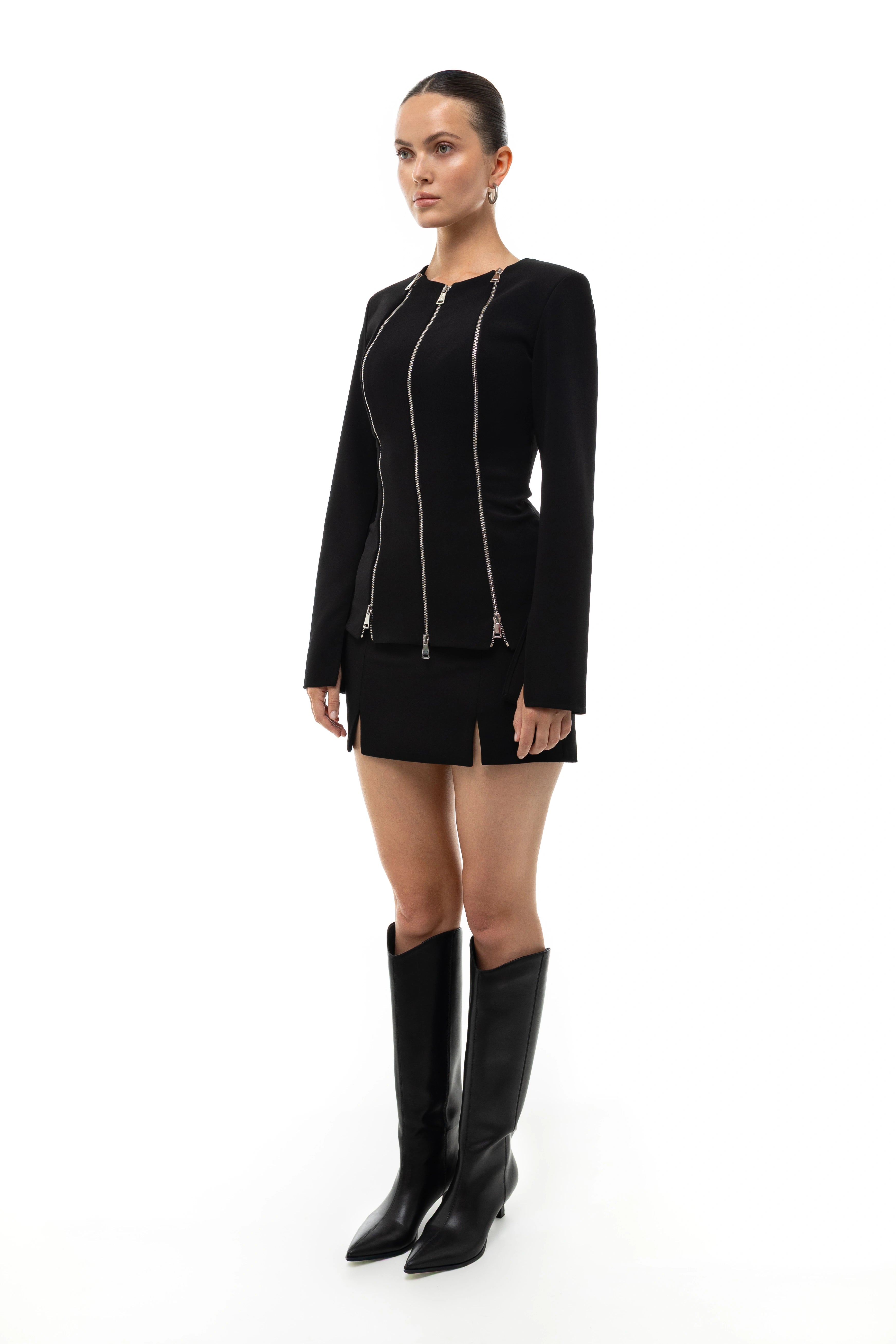 Woman wearing a black outfit with zippers on a white background. Blazer. Mini skirt. Boots. Soluje