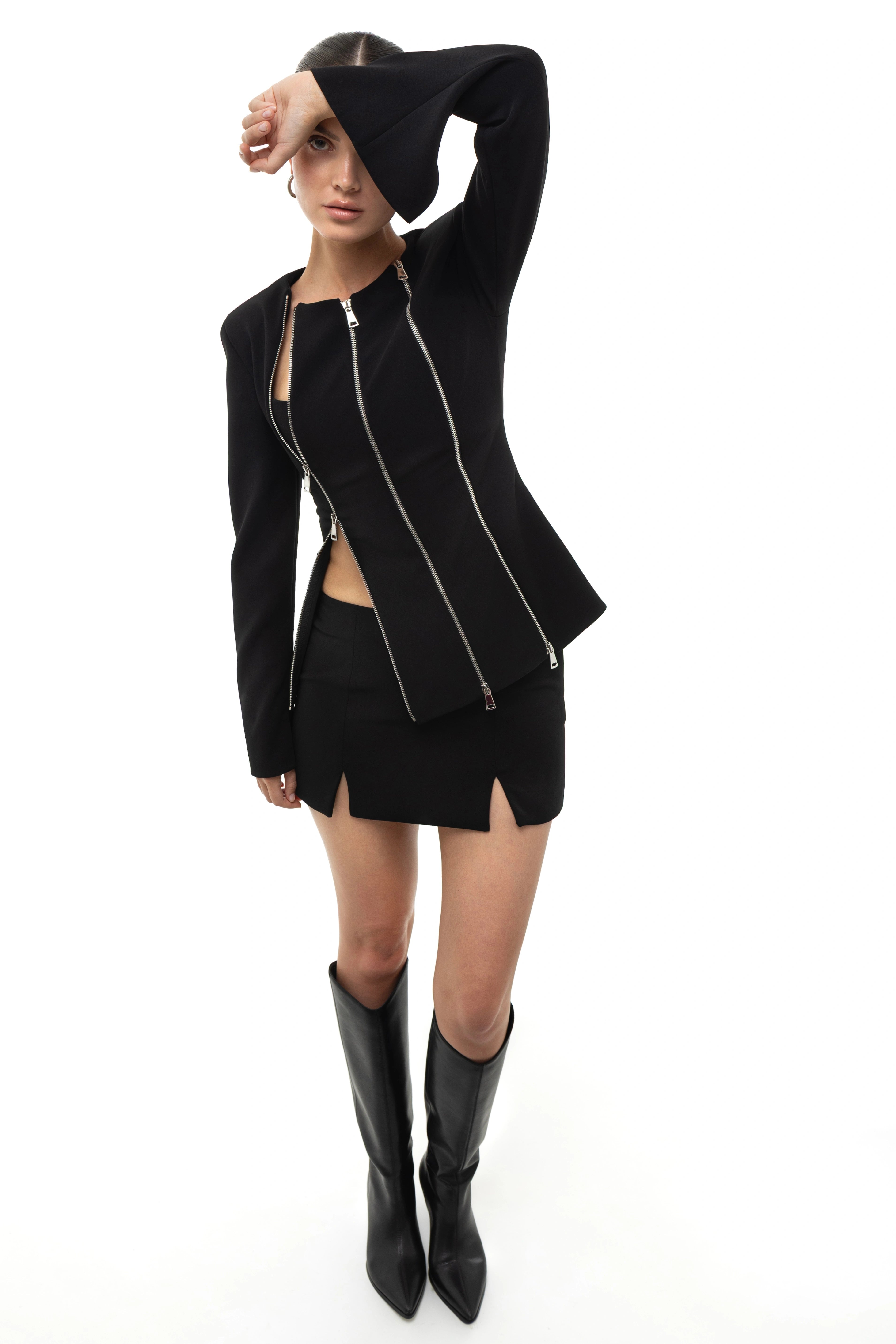 Woman wearing a black outfit with a jacket with zippers, skirt, and boots on a white background. Soluje