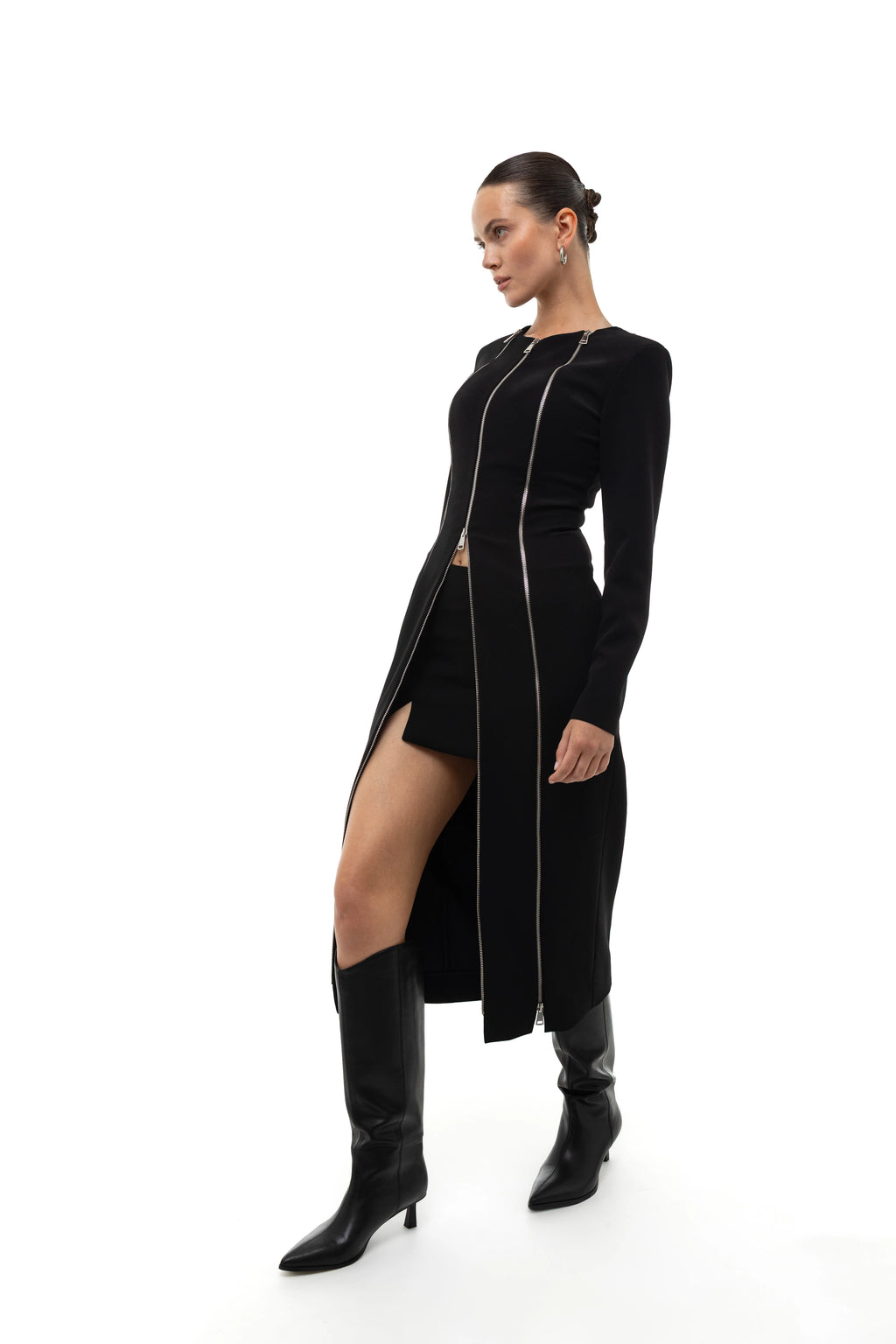 Woman wearing a long black coat and dress with a high slit decorated with zippers, mini skirt and matching knee-high boots on a white background