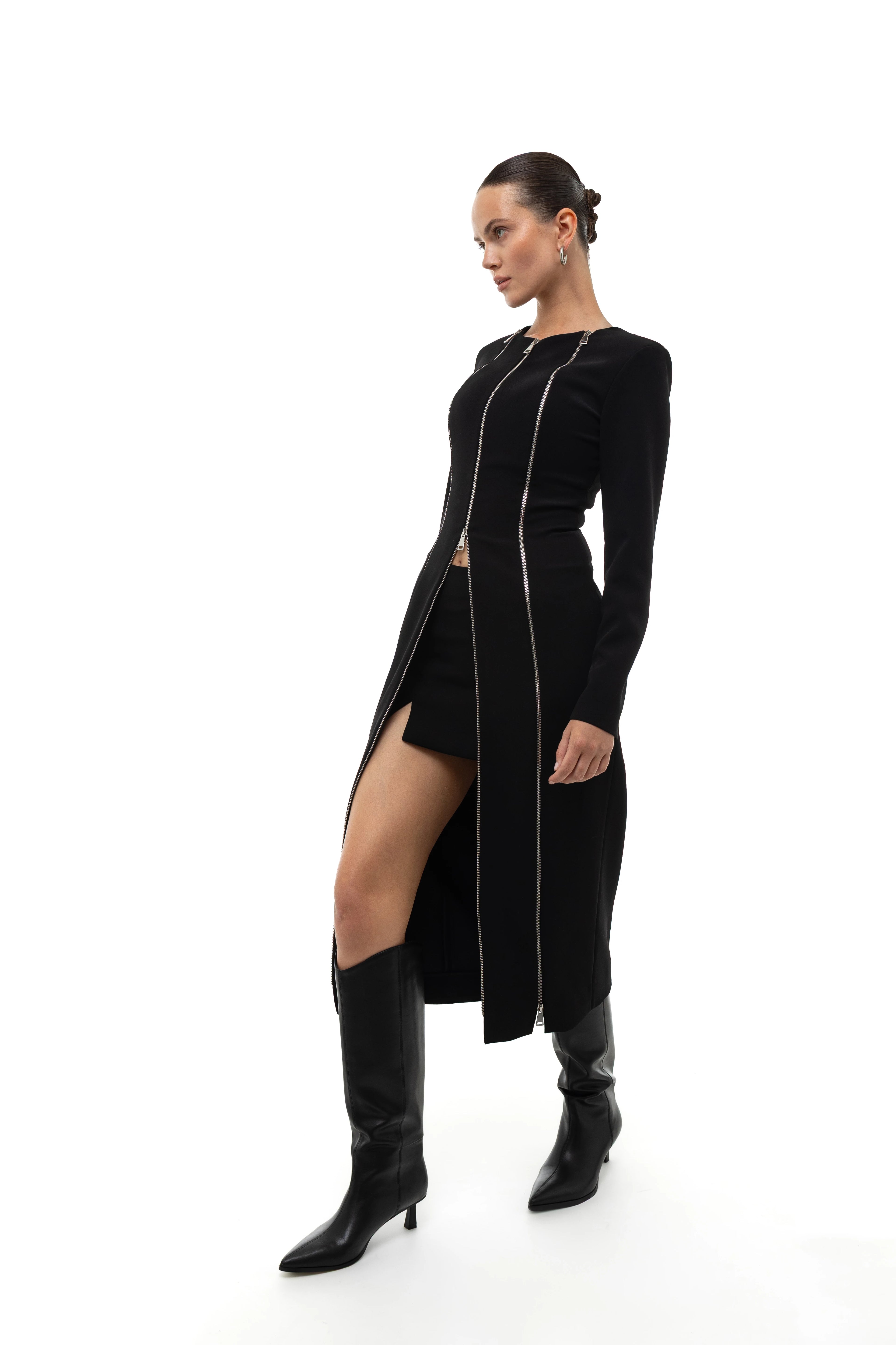 Woman wearing a long black coat and dress with a high slit decorated with zippers, mini skirt and matching knee-high boots on a white background
